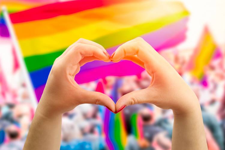 person making heart shape with hands with rainbow flags behind them for a pride celebration